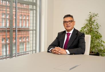 Vijay Bange in a dark suit, white shirt and red tie sits at an empty table with windows behind him showing a red brick building.