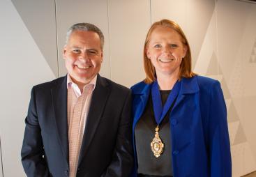 A smiling man and woman in business attire (dark suit, open necked shirt, blue jacket, black top) look at the camera