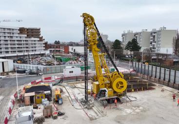 An elevated view of a yellow Soilmec SC-110 Urban cutter working on the Grand Paris Express, constructing 1,200mm-thick diaphragm walls up to 38 metres deep at Rueil–Suresnes Mont-Valérien station, surrounded by houses, low rise buildings and roads.