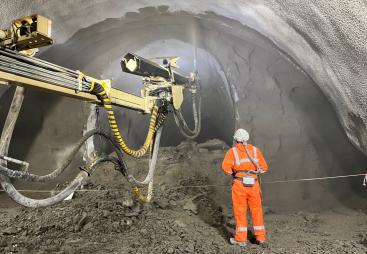 A construction worker in orange PPE uses a remote control unit to control a Normet concrete sprayer in a tunnel