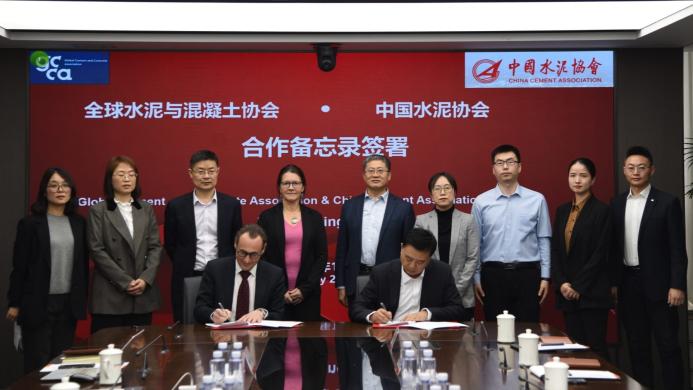 Several people standing around a table looking at two men signing a document