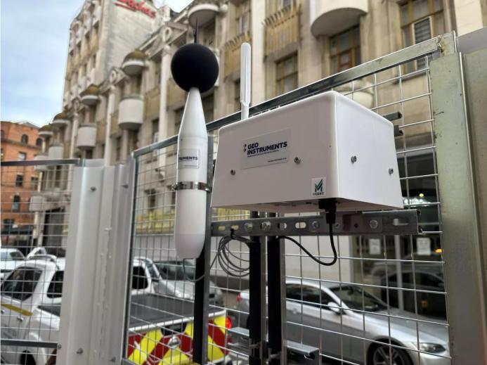 a dust monitoring station attached to a metal fence withan building in the background