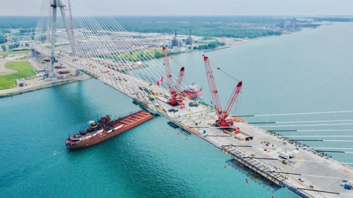 Aerial view of a bridge under construction across a river with two red cranes on its deck