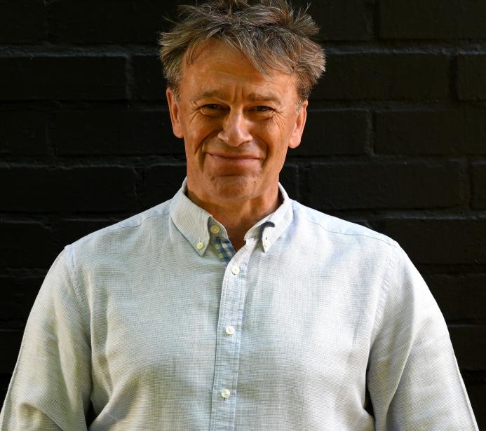 A head and shoulders picture of a man with unruly hair in a white shirt against a black background