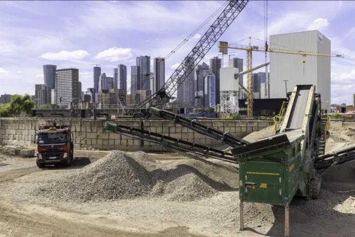 Piles of aggregate in a yard with a truck and green screen with a crane and tall buildings in the background
