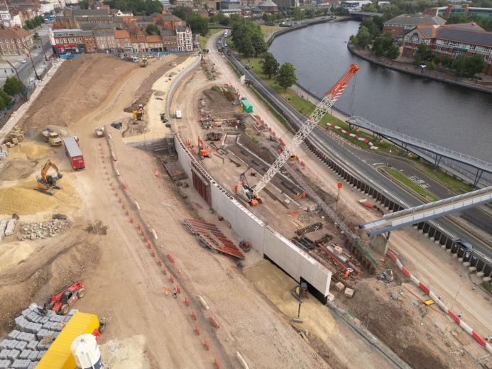 A large construction site by a river, including an orange crawler crane and other construction equipment