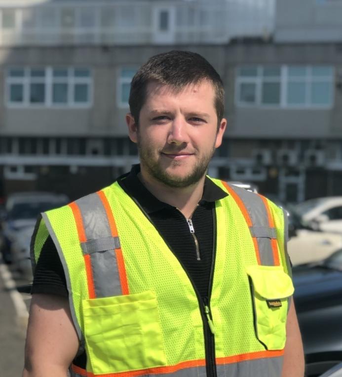 A man with short dark hair wearing yellow PPE in front of a mid-rise building