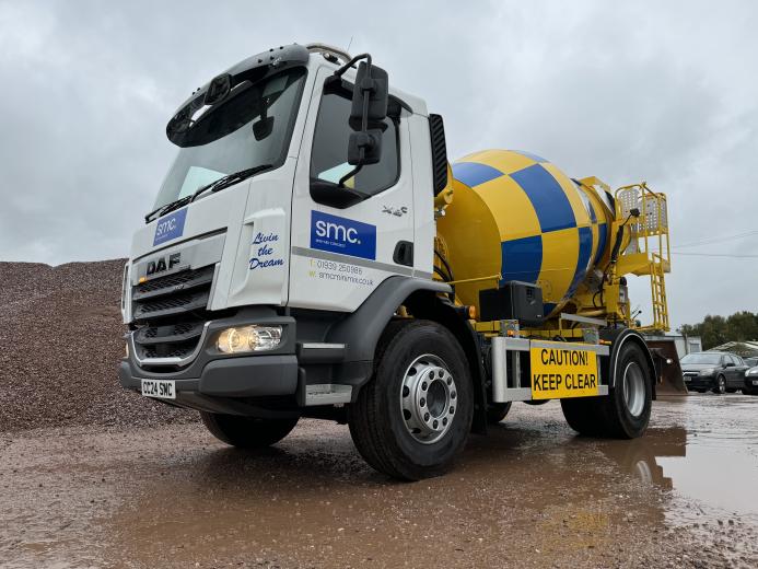 A white, yellow and blue concrete mixer truck in a builder's yard