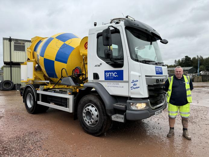 A man in yellow PPE stands in front of a white truck fitted with a yellow and blue concrete mixer