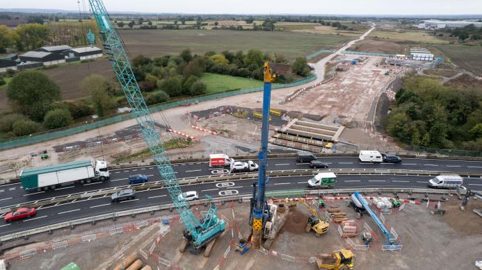 A green crane and yellow piling rig photographed from the air installing piling on a large construction site by a dual carriageway