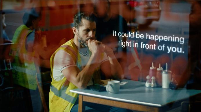 A man with dark hair in a top knot, wearing yellow PPE sitting at a cafe table looking anxious