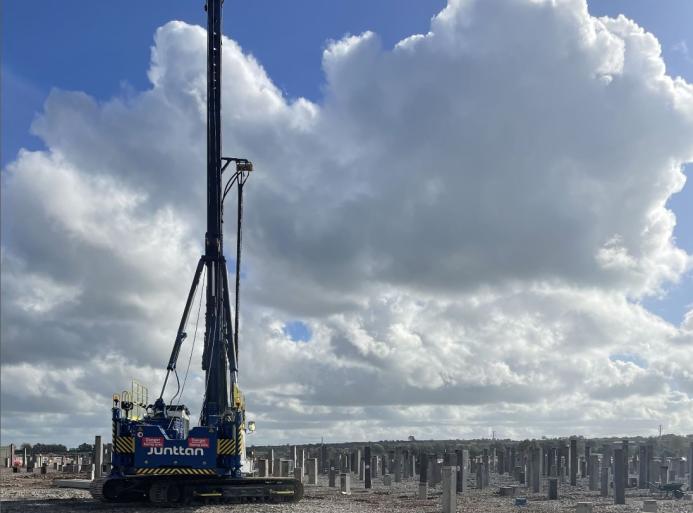 A Junttan piling rig stands in front of hundreds of pile caps, with a blue sky and dramatic clouds