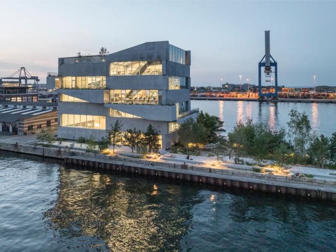 A seven storey concrete building next to a body of water with a ship loading crane in the background