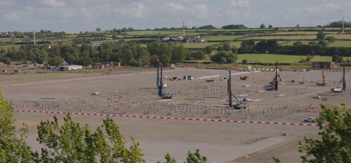 An aerial view of piling rigs working on a huge, level construction site surrounded by trees and fields