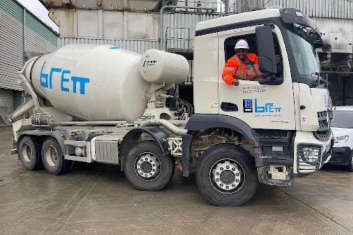 A white concrete mixer truck being driven by a man in red PPE