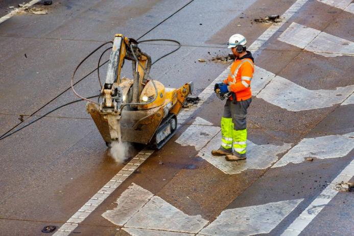 A man in orange, grey and yellow PPE uses a remote controlled piece of construction equipment fitted with a breaker to carry out repairs to a concrete road