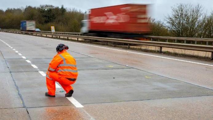 A man in orange PPE carrying out an inspection of an empty concrete road, while a red truck passes in the background on the opposite carriageway