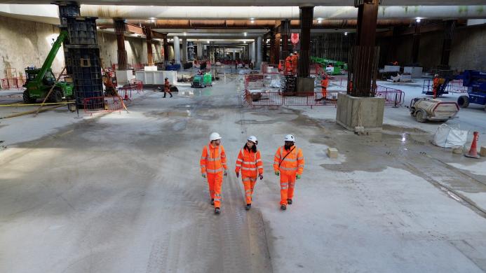 Three people in orange PPE walk across the competed base slab in the 850m long underground station box at Old Oak Common.
