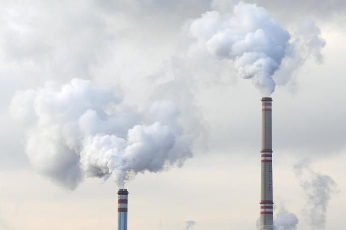 The top of two tall chimneys against a grey sky billow "smoke" into the atmosphere