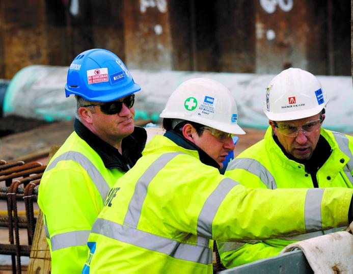 Three Costain construction workers in yellow PPE look at plans on a construction site.