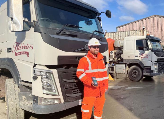 A man in orange PPE standing in front of a cement mixer holding a mobile device that uses the Podfather app to access their schedules and delivery notes, navigate routes, and record delivery details digitally. 