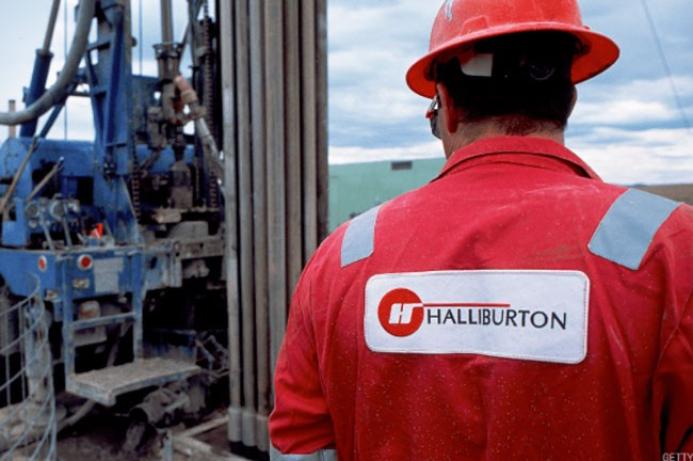 A man in red overalls and red hard hat stands with his back to the camera, a Halliburton logo on his back, surveying a Halliburton Energy Services project.