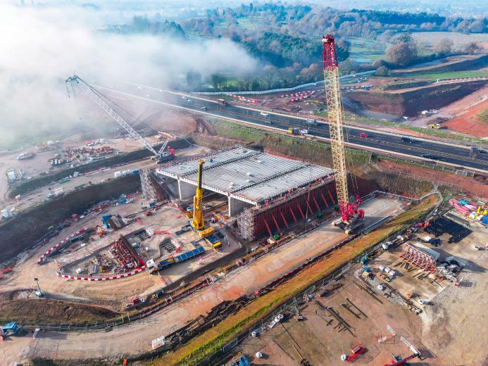 An aerial view of the A46 Kenilworth Bypass box structure under construction with a large yellow and red crawler crane and a smaller white telescopic crane and white crawler crane. Image: HS2.