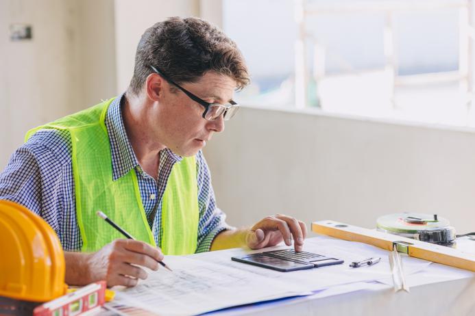 A man in glasses, blue shirt and yellow PPE vest sits working at a desk.