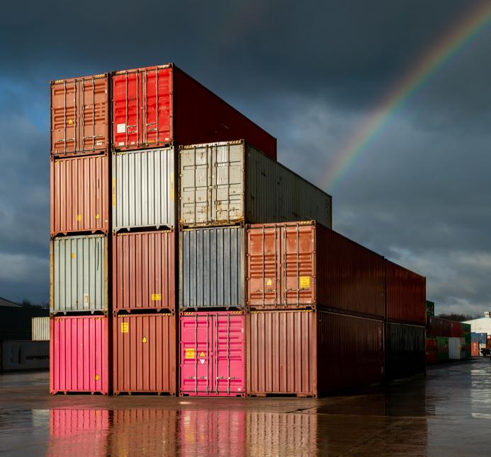 Multi-coloured containers stacked up in a yard with storm clouds and a rainbow in the background