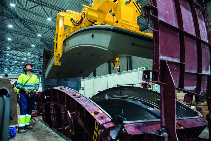 A yellow and blue PPE clad worker in a factory uses a mould and lifting device for precast tunnel segments, France, CBE