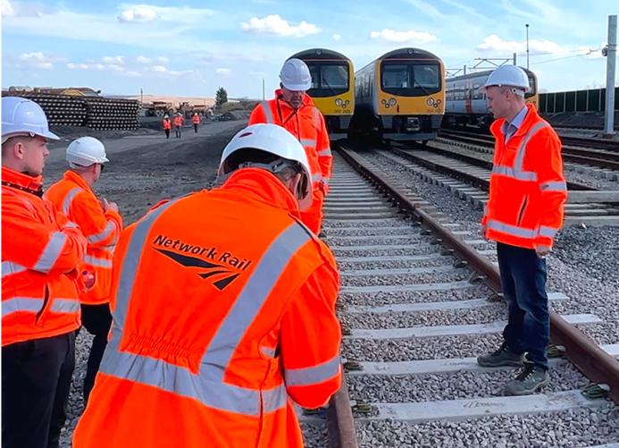 A group of people in Network Rail branded PPE inspect a concrete railway sleeper. The first-generation low-carbon concrete sleepers were installed by Concretene at GCRE in South Wales using 3.75 kg less cement per unit.