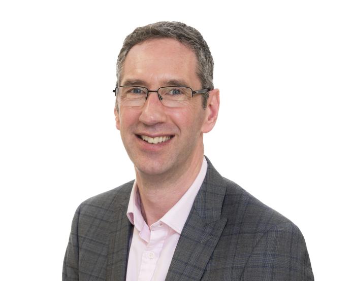 A head and shoulders shot of Peter Reekie of the Scottish Futures Trust will speak on sustainable infrastructure at the Glasgow Industry Day wearing glasses, an open necked pick shirt and a grey over-check jacket