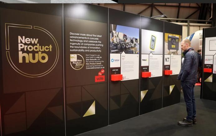 A man stands right of frame looking at entries to the UK Concrete Show's New Product Hub, which are displayed on a large, floor standing board.
