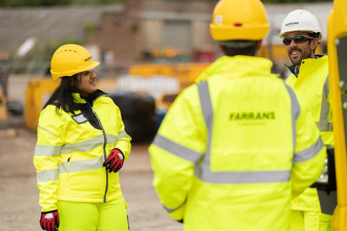 Three people in yellow PPE stand talking with construction machinery in the background