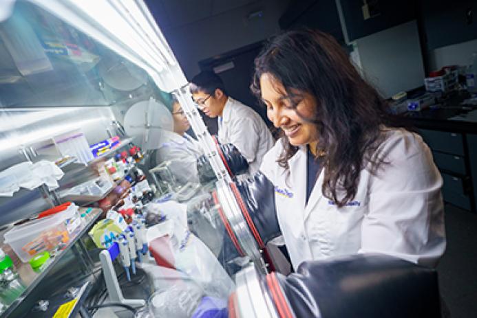Two people working in a laboratory, one in the background, one in the foreground in front of a glass fronted cabinet full of scientific instruments.
