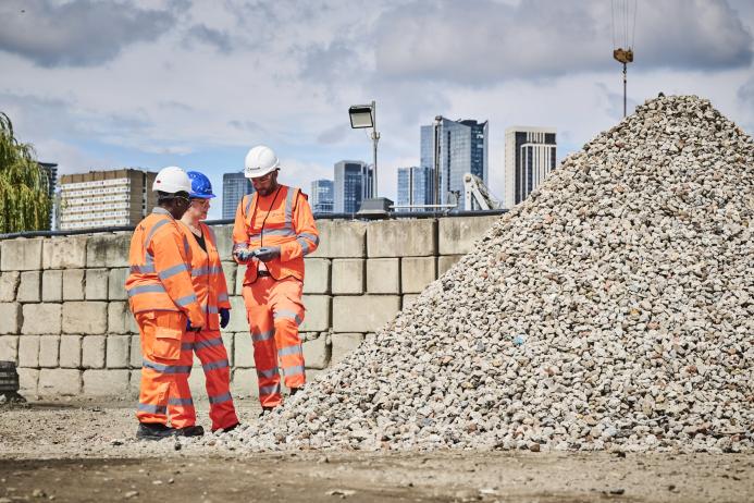 Three people in orange PPE stand discussing aggregate needs in a construction materials yard with a wall and city scape in the background