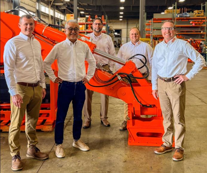 Five men dressed in smart shirts and trousers stand around a large red excavator arm