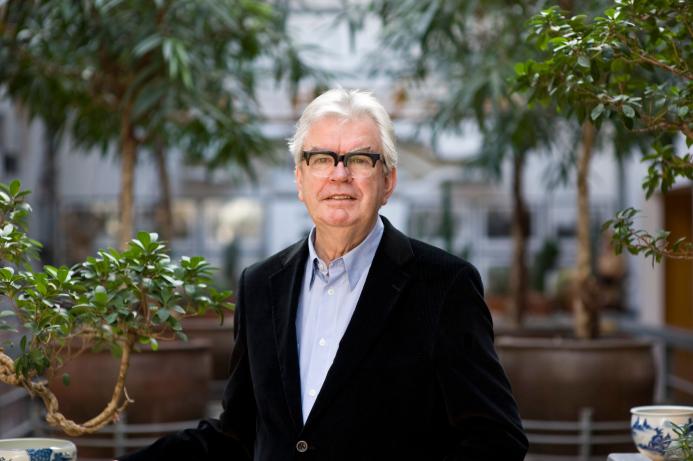 A grey-haired Sir Terry Farrell (1938-2025) wearing glasses, a dark jacket and light blue shirt photographed in front of an avenue of small trees in an urban setting © richard gleed / farrell