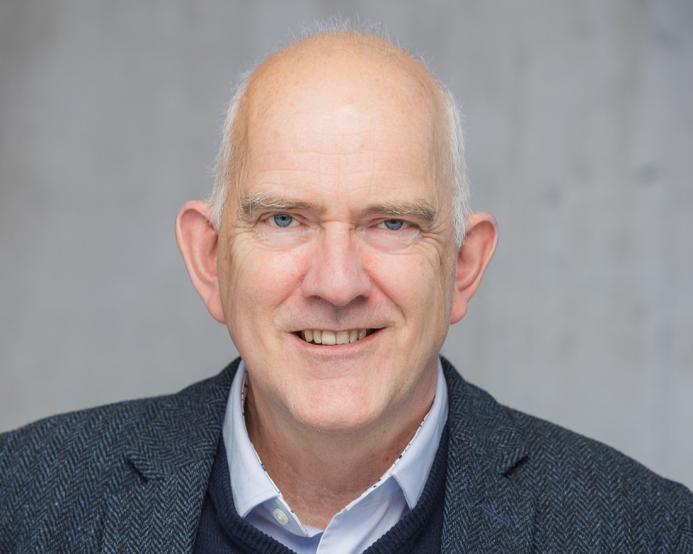 Head and shoulders photo of a bald, smiling man in open necked shirt, jumper and jacket