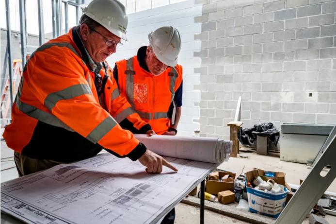 Two construction workers look at plans laid out on a table inside an under construction building