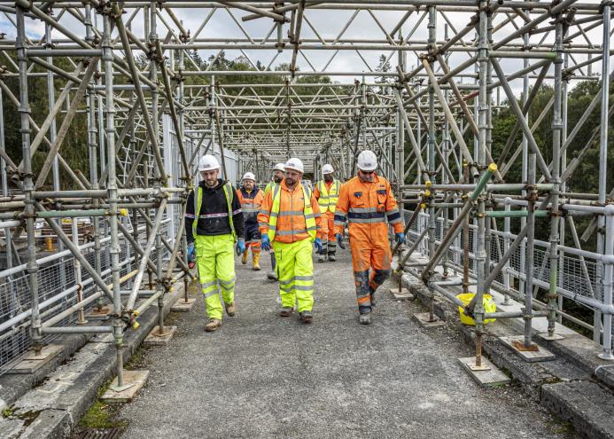 A group of construction workers walk along a concrete walkway, through a "tunnel" of scaffolding, wearing a variety of PPE , including orange hi vis jackets, yellow trousers, white hard hats and eye protectors