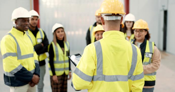 A group of youg people wearing yellow PPE and white and yellow hard hats listen to an older worker