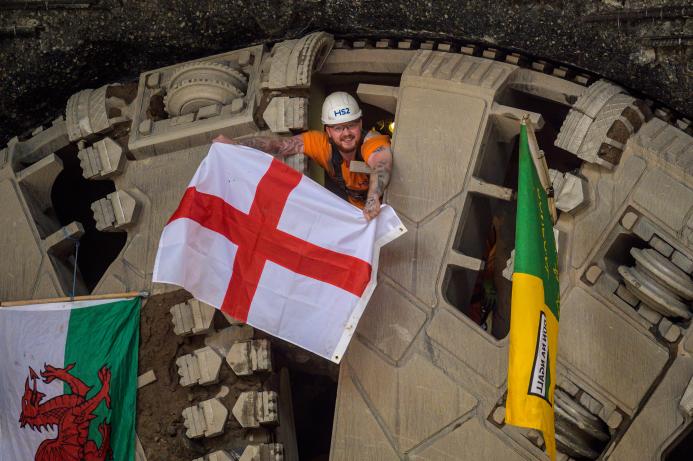 A man wearing a hard hat and waving a Cross of St George flag pokes his head through a TBM cutter wheel with a Welsh flag and another unidentified flag adorning the cutter head as HS2 engineers celebrate the breakthrough of tunnel boring machine Elizabeth at Washwood Heath in Birmingham, completing the 3.5-mile Bromford tunnel under the M6. (Image © HS2 Ltd)