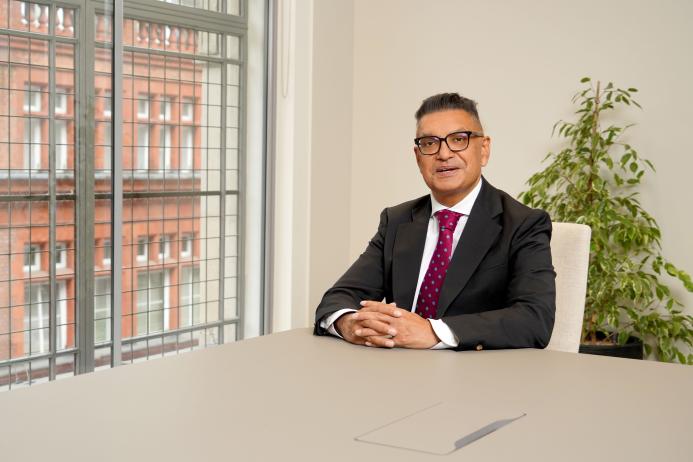 Vijay Bange in a dark suit, white shirt and red tie sits at an empty table with windows behind him showing a red brick building.