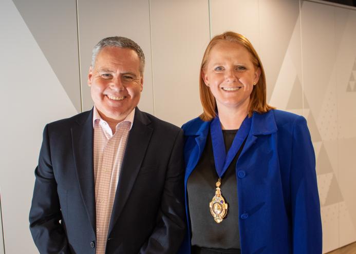 A smiling man and woman in business attire (dark suit, open necked shirt, blue jacket, black top) look at the camera
