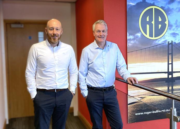 Two middle-aged men in smart, business casual wear stand on a stair landing in front of a large photo