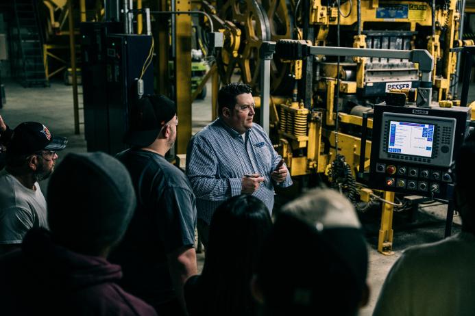 A bearded man, surrounded by a small group of people all looking at an LCD screen in a factory surrounded by heavy equipment