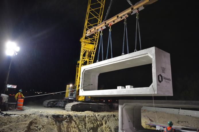 A yellow Kobelco crawler crane lifts a precast, rectangular concrete element