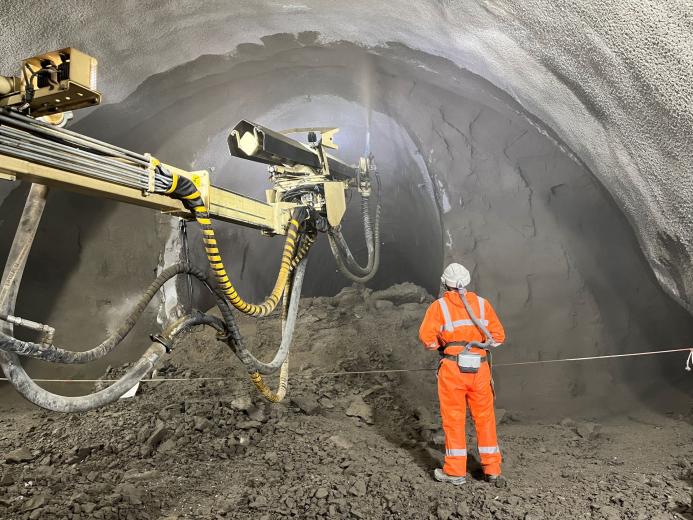 A construction worker in orange PPE uses a remote control unit to control a Normet concrete sprayer in a tunnel