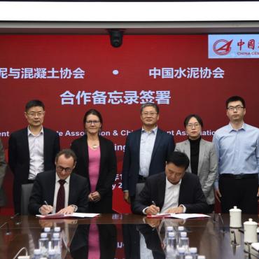 Several people standing around a table looking at two men signing a document
